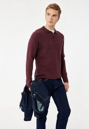 Jeune homme portant une chemise à manches longues bordeaux et un jean foncé, tenant une veste en denim pliée, regardant sur le côté contre un fond blanc.