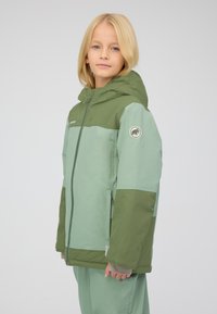 Giovane con capelli biondi della lunghezza delle spalle, che indossa una giacca con cappuccio verde chiaro e scuro e pantaloni abbinati, in piedi davanti a uno sfondo neutro.