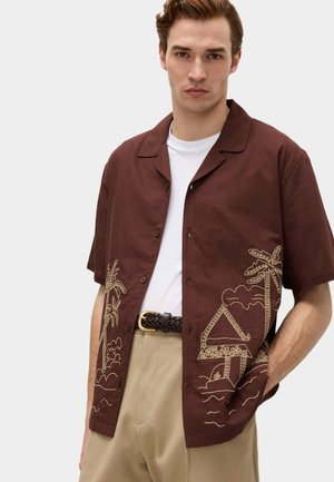 Ung mand iført en brun kortærmet skjorte med beige palme- og husbroderi over en hvid t-shirt og beige bukser.