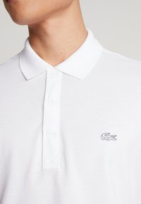 Polo blanc en coton texturé. Il présente un col classique, une patte de boutonnage et un petit logo crocodile brodé sur la poitrine.