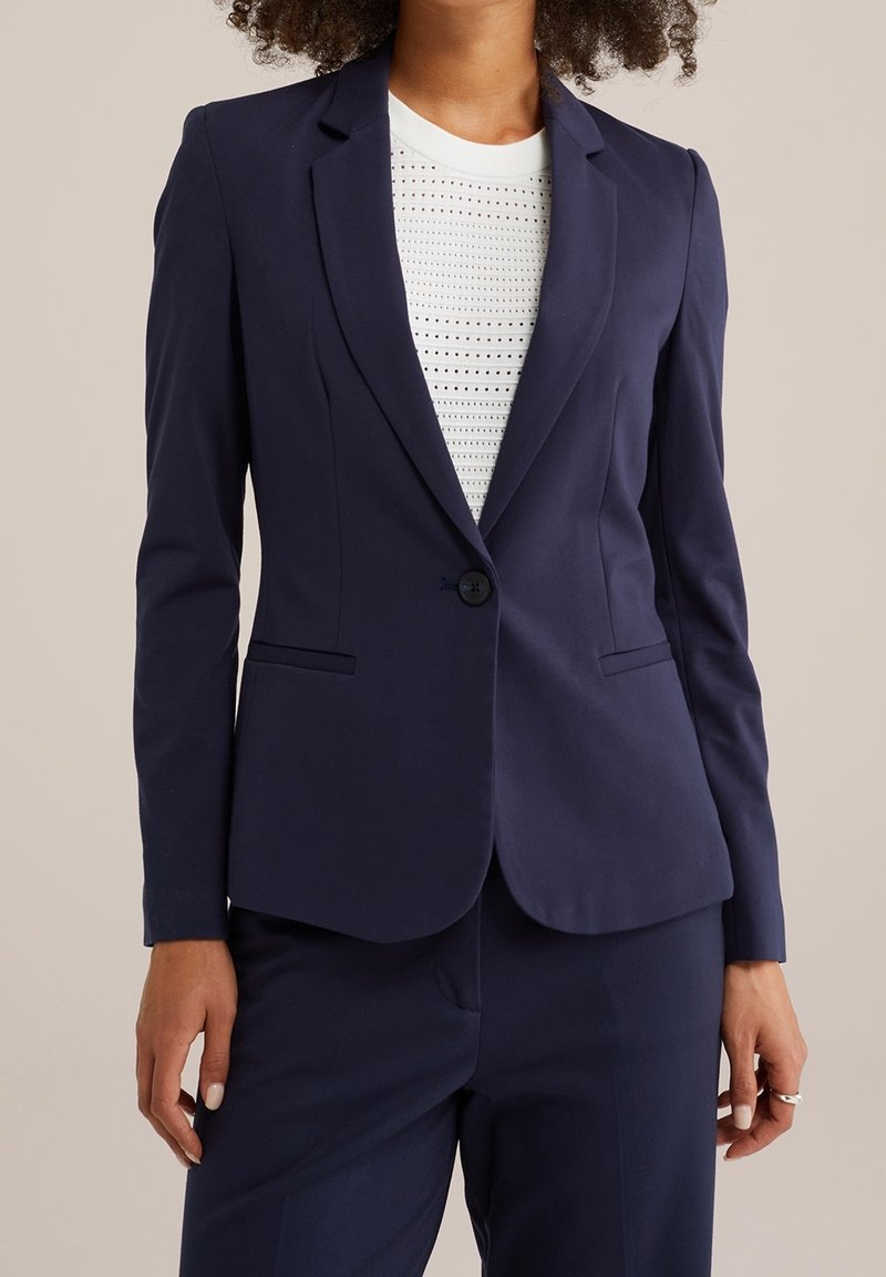 Woman wearing a navy blazer and matching pants with a white perforated top, standing against a plain beige background.