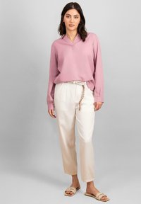 Blouse à manches longues en rose doux, avec un col en V et une coupe décontractée, associée à un pantalon blanc et une ceinture tressée. Sandales avec un détail tissé.