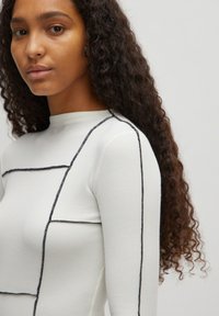 Witte geribde aansluitende top met hoge halslijn, voorzien van zwarte contraststiksels in geometrische patronen. Lange mouwen en een zachte textuur.