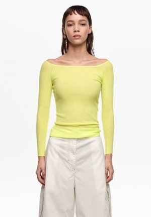 Joven mujer que lleva una blusa de manga larga y cuello caído de color amarillo neón y pantalones blancos sueltos de talle alto, de pie frente a un fondo liso.