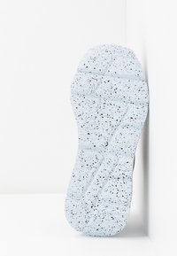 Semelle d'une chaussure blanche avec un motif moucheté noir reposant verticalement contre une surface d'angle blanche.