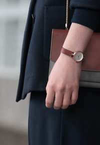 Montre à bracelet en cuir marron avec un cadran rond blanc et des accents dorés, portée sur une main tenant un sac à main marron et gris.