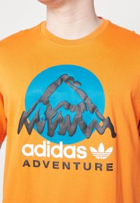 Oranžové bavlněné tričko s modrým kruhovým motivem horského designu, černým obrysem a bílým nápisem "adidas ADVENTURE" pod ním.