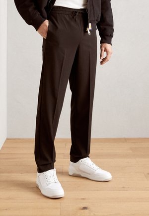 Pantalons de survêtement noirs avec taille élastique et cordon de serrage, dotés de poches latérales, associés à des baskets blanches sur un sol en bois.