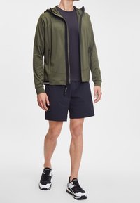 Felpa con zip verde oliva, con cappuccio, maglietta nera a maniche corte sotto, pantaloni corti neri e sneaker nere e bianche. Tessuto morbido con due tasche laterali.