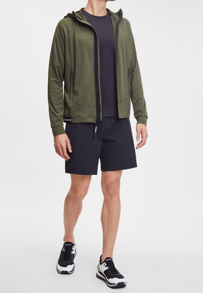Felpa con zip verde oliva, con cappuccio, maglietta nera a maniche corte sotto, pantaloni corti neri e sneaker nere e bianche. Tessuto morbido con due tasche laterali.