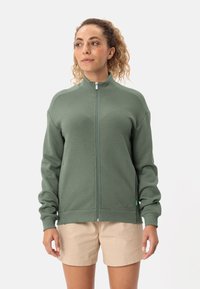 Giacca verde con zip caratterizzata da un motivo texturizzato, colletto alto e polsini a coste, abbinata a pantaloni corti beige.
