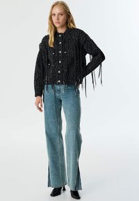 Chaqueta negra texturizada con flecos, botones plateados y un diseño corto; combinada con unos vaqueros de pierna ancha azul claro que presentan cremalleras laterales.