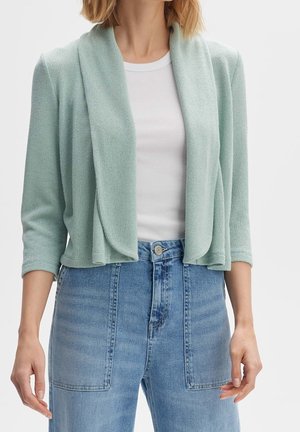 Femme portant un cardigan vert clair ouvert devant avec des manches trois-quarts sur un top blanc côtelé et un jean bleu taille haute.