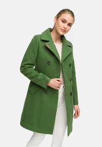 Manteau double boutonnage en mélange de laine vert avec grand col cranté, deux poches avant et boutons noirs. Détail en cuir sur la manche.
