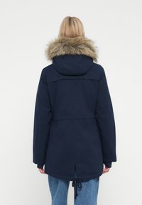 Donna che indossa una parka blu navy con cappuccio bordato di pelliccia, jeans blu, di spalle, su uno sfondo bianco uniforme.