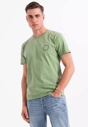 Camiseta verde de manga corta hecha de tela suave, con un cuello redondeado y manchas negras dispersas. Incluye un logo circular en el pecho.