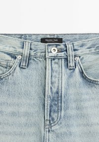 Jean en denim bleu clair avec fermeture à boutons et passants pour ceinture, portant une étiquette noire indiquant "Massimo Dutti Made in Turkey" à la taille.