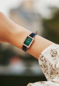 Reloj rectangular con una esfera verde, acentos dorados y adornos parecidos a diamantes, con una correa de cuero negra.
