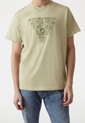 Beżowa koszulka z krótkim rękawem z zielonym trójkątnym logo GUESS i motywem liści, noszona przez stojącą osobę w jasnoniebieskich dżinsach.