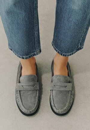 Feet wearing grey suede loafers paired with blue denim jeans standing on a light grey floor.
