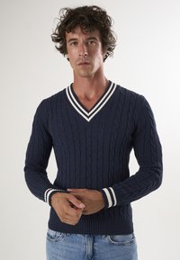 Maglione lavorato a maglia blu navy con scollatura a V, caratterizzato da motivi a treccia e polsini e orlo a righe bianche. Design aderente.