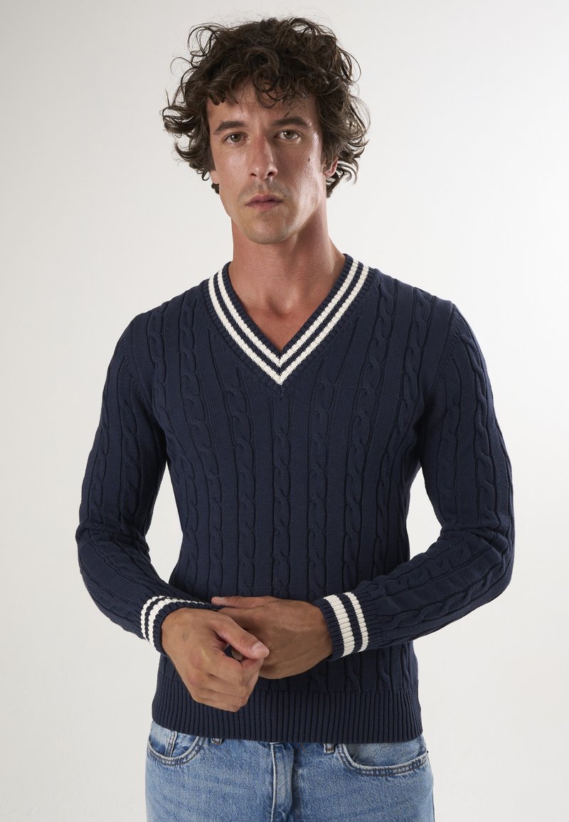 Maglione lavorato a maglia blu navy con scollatura a V, caratterizzato da motivi a treccia e polsini e orlo a righe bianche. Design aderente.