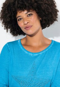 Türkises Langarmshirt mit einem großen Sternmuster aus metallischen Nieten. Weicher Stoff mit rundem Ausschnitt und lässiger Passform.