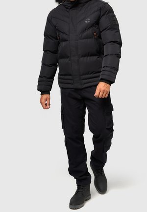 Zwarte pufferjas met een rits aan de voorkant, horizontale stiksels en een capuchon; gecombineerd met zwarte cargobroeken en zwarte veterschoenen.