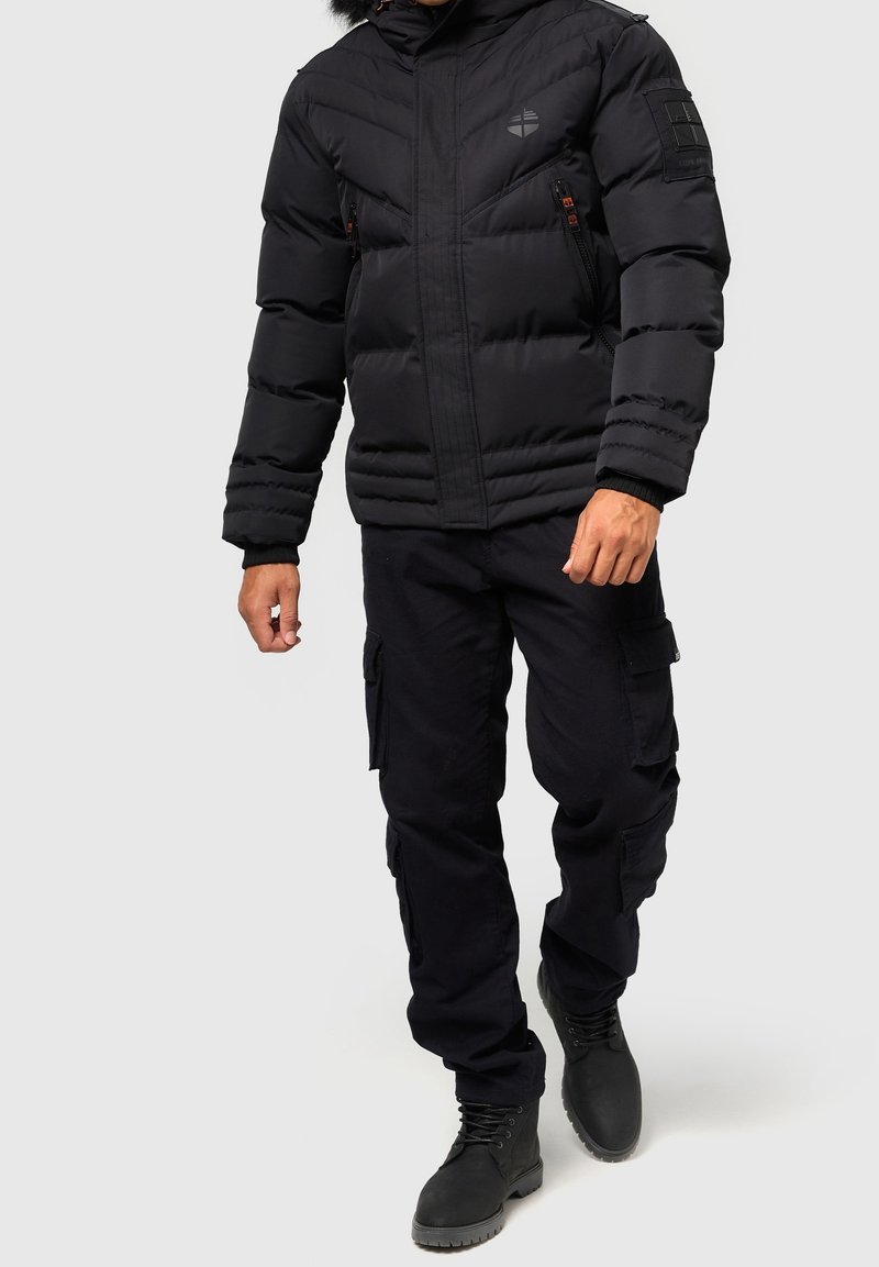 Zwarte pufferjas met een rits aan de voorkant, horizontale stiksels en een capuchon; gecombineerd met zwarte cargobroeken en zwarte veterschoenen.