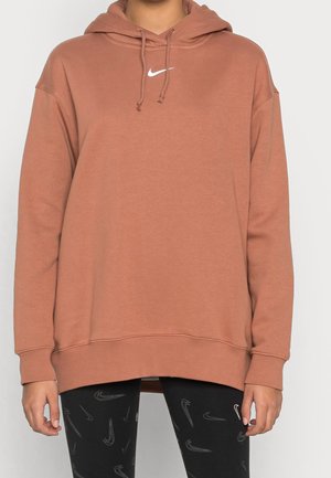 Osoba nosząca brązową bluzę z kapturem Nike z białym logo i czarne legginsy z wzorem z wieloma logo Nike swoosh.