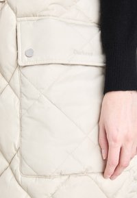 Colete bege acolchoado com um bolso frontal de fecho. Apresenta o logótipo bordado "Barbour". Tecido suave com uma textura fina e acolchoada.