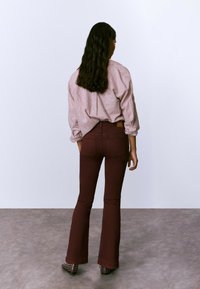 Mujer con pelo largo de espaldas, vistiendo una camisa de rayas rosas, pantalones acampanados marrones y zapatos negros con tachuelas sobre un suelo gris.