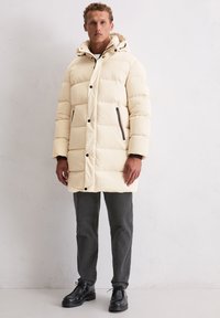 Crèmekleurig pufferjack met horizontale doorgestikte secties, zwarte ritssluitingen en een capuchon. Gecombineerd met donkere textuurbroeken en zwarte schoenen.