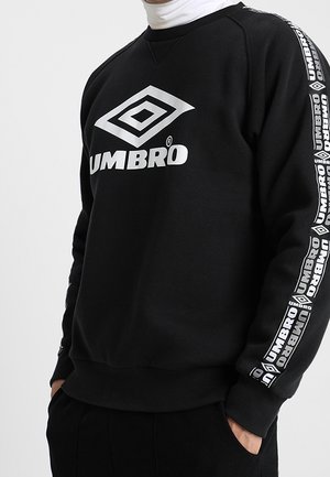 Schwarzer Sweatshirt mit weißem Umbro-Logo auf der Brust und wiederholtem Umbro-Branding an den Ärmeln, getragen über einem weißen Rollkragen.