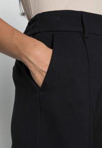 Svarta byxor med ett mjukt, elastiskt tyg; veckad design; och ett elastiskt midjeband med en sidoficka synlig.