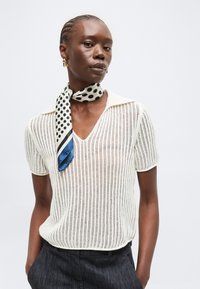 Γυναίκα με πλεξούδες, φοράει λευκό πλεκτό μπλουζάκι με κοντά μανίκια, μαύρο παντελόνι και φουλάρι με πουά δεμένο γύρω από το λαιμό της.