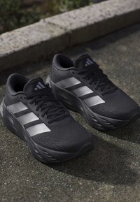 Ζευγάρι μαύρα παπούτσια τρεξίματος Adidas με τρεις ασημένιες ρίγες, μαύρα κορδόνια και υφασμένους ενδιάμεσους πάτους σε άσφαλτο κοντά σε λίθινη άκρη.