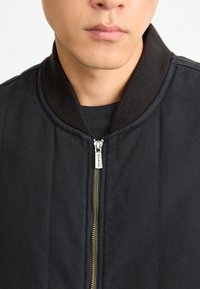 Črna bomberska jakna z rebrenim ovratnikom, zlatim zadrgo in prešito teksturo. Podrobno osredotočanje na podrobnosti zadrge in ovratnika.