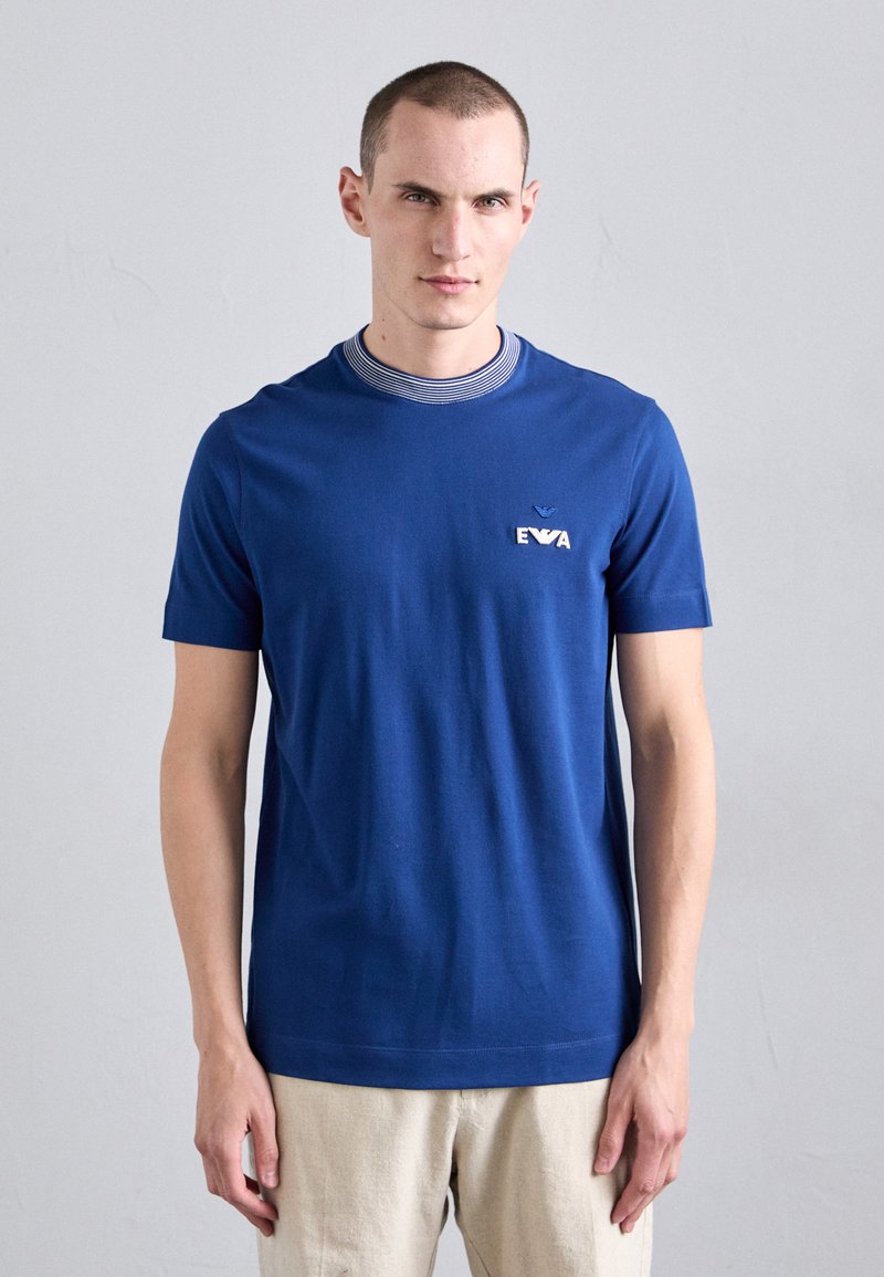 T-shirt en coton bleu à manches courtes, avec un col côtelé orné de rayures bleu marine et blanches, et un petit logo brodé blanc sur la poitrine.