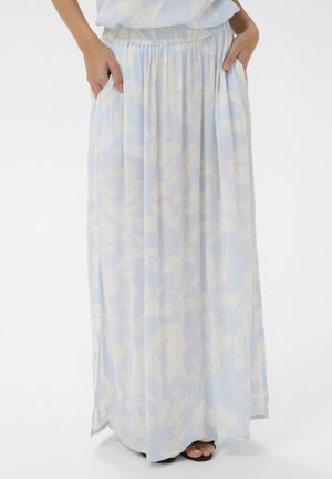 Femme portant une longue jupe bleu clair à motif floral blanc, les mains dans les poches, montrant ses pieds en sandales noires sur fond blanc.