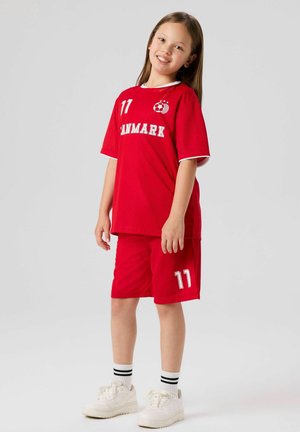 Fille souriante aux longs cheveux portant un maillot et un short de football rouge du Danemark avec le numéro 11, des baskets blanches et des chaussettes rayées sur un fond uni.