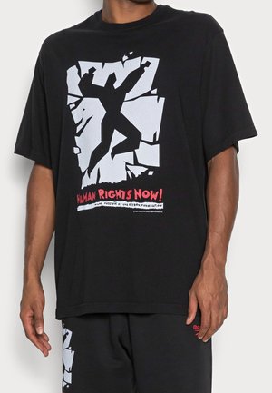 T-shirt en coton noir avec un design graphique audacieux en argent, représentant une silhouette en train de briser un mur, avec un texte rouge disant "DROITS DE L'HOMME MAINTENANT !"