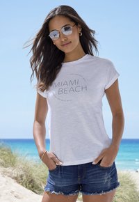 White relaxed-fit t-shirt with "MIAMI BEACH" text, paired with dark blue denim shorts. The setting features a beach background with blue skies.