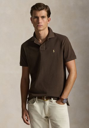 Młody mężczyzna ubrany w ciemnobrązową koszulkę polo z logo, jasnobeżowe spodnie, brązowy pasek i zegarek na rękę z niebieską tarczą, stoi z jedną ręką w kieszeni.