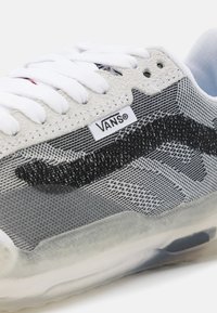 Tênis Vans com parte superior em malha cinza, detalhes em preto, atacadores brancos e sola translúcida. Etiqueta do logo visível na lateral.