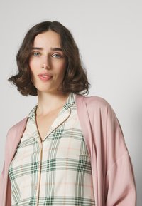 Ein leuchtend rosa, fließender Morgenmantel über einem cremefarbigen Karo-Oberteil, das grüne und pfirsichfarbene Nuancen aufweist. Der Stoff wirkt weich, mit einer lockeren Passform und Knopfverschluss.