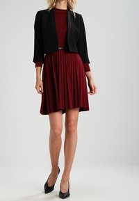 Chaqueta negra corta sobre un vestido burdeos con pliegues y un corpiño ajustado. El vestido tiene mangas largas y la chaqueta presenta un cuello de satén.