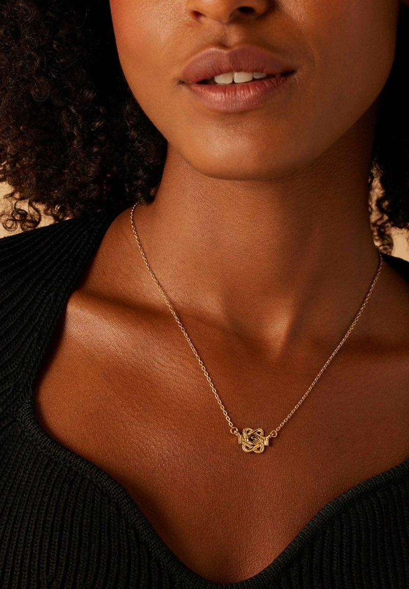 Collier en or avec un design de nœud complexe, associé à un haut noir texturé, mettant en valeur le teint de la peau et les détails de fabrication.