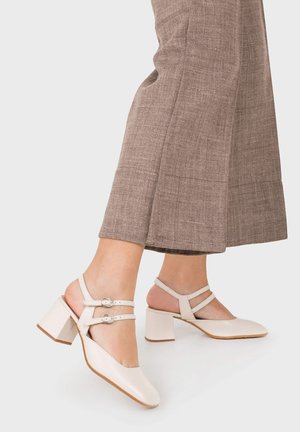 Mujer con zapatos beige de tacón grueso con doble correa, combinados con pantalones cortos marrones con textura sobre un fondo liso.