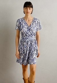 Robe portefeuille florale en blanc et marine, avec des manches courtes à volants, un décolleté en V et un lien à la taille. Texture douce avec une coupe ample.